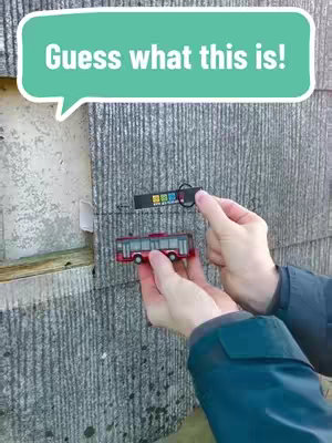 A person holds a small red bus model and a black tag/keychain in front of a gray stone wall. A speech bubble on top reads, "Guess what this is!"