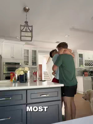 Two young men hugging in a kitchen. One is wearing a green shirt and black shorts, the other a white t-shirt and red shorts. The kitchen has white cabinets, a grey island, and a dog can be seen near the edge.