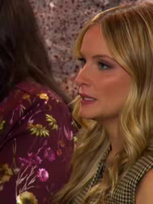 The image shows a profile view of a woman with blonde wavy hair and fair skin. She is facing right with a slight smile, and appears to be in conversation. Part of another person is visible on the left side of the frame. The background is out of focus and suggests an indoor setting.