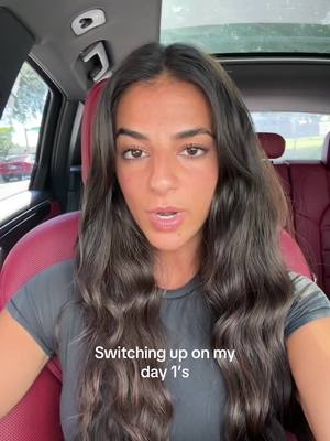 A woman is in a car, likely filming a selfie-style video. She has long, wavy dark hair and is wearing a gray t-shirt. The background shows the interior of the car, including the seat and side window. Text overlay provides the video's hook.
