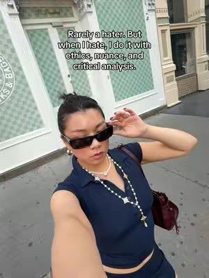 A selfie-style shot featuring a woman holding the camera, looking towards the viewer. She's wearing dark sunglasses, a dark blue top, and a pearl necklace with a star charm. The background shows a city street with buildings and shop fronts.