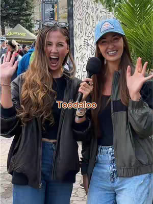 Two young women stand outdoors, smiling and waving at the camera. The woman on the left is wearing a black top and jacket with her mouth open. The woman on the right has a blue Red Bull hat and also holds a microphone. Text that says "Toodaloo" is overlaid on the image.