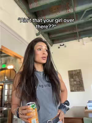 A young woman stands indoors, holding a can and looking up and slightly off-camera with a surprised look. She has long dark hair, wearing a gray tank top, a silver necklace and bracelets.  The background includes an interior with architectural detail, wooden beams and a visible sign.