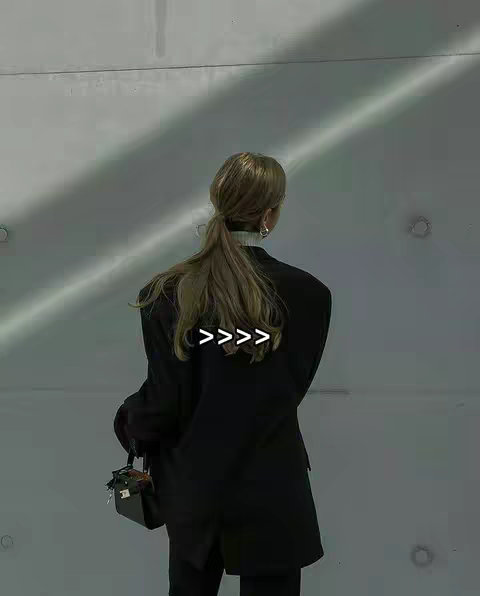 A woman stands with her back to the camera in a minimal, concrete environment. She is wearing a black blazer and holding what appears to be a small dark purse. Her blonde hair is pulled back in a ponytail, and the image has a muted, shadowy lighting with angled light beams creating shadows on the wall.