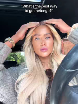 A woman with long blonde hair and light blue eyes sits in a car, looking forward with her hands raised towards her head. She is wearing a gold wristwatch and a gray sweater. The background shows a car interior.