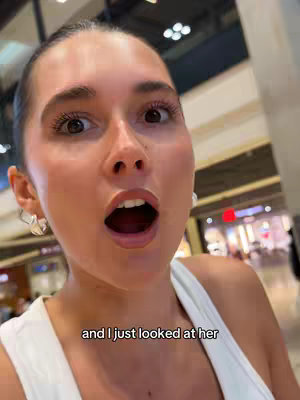 A close-up shot of a woman with wide eyes and an open mouth, conveying surprise or shock. The background is blurred, but suggests an indoor setting with lights. The woman is wearing a white top and earrings. The text overlay reads, "and i just looked at her."