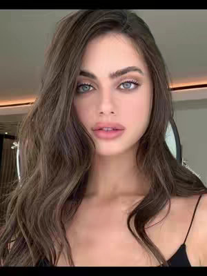 A close-up portrait of a young woman with long, wavy brown hair, light skin, and striking blue eyes. She gazes directly at the camera with a soft, inviting expression. Her lips are gently parted, suggesting a smile. She wears subtle makeup, enhancing her natural features. The background is subtly blurred, focusing attention on the woman. She is wearing a black spaghetti strap top.
