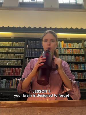 A woman is holding a large, dark maroon water bottle to her lips while posing in an old library filled with books. The text overlay "LESSON 1: your brain is designed to forget" appears in white text at the bottom of the image.