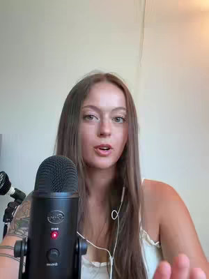 A woman with long hair is centered in the frame, looking directly at the camera. She is holding a microphone with her left hand and has a tattoo on her left arm. The background is a plain wall.