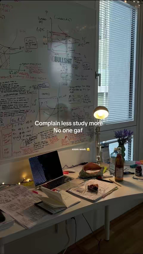 The image shows a desk setup in front of a window with blinds. A whiteboard covered in handwritten notes and diagrams is visible on the wall. On the desk are a laptop, books, papers, a lamp, and miscellaneous items such as a bottle of juice, snacks, and a small bouquet of flowers. The text overlay reads: "Complain less study more. No one gaf. exam week"