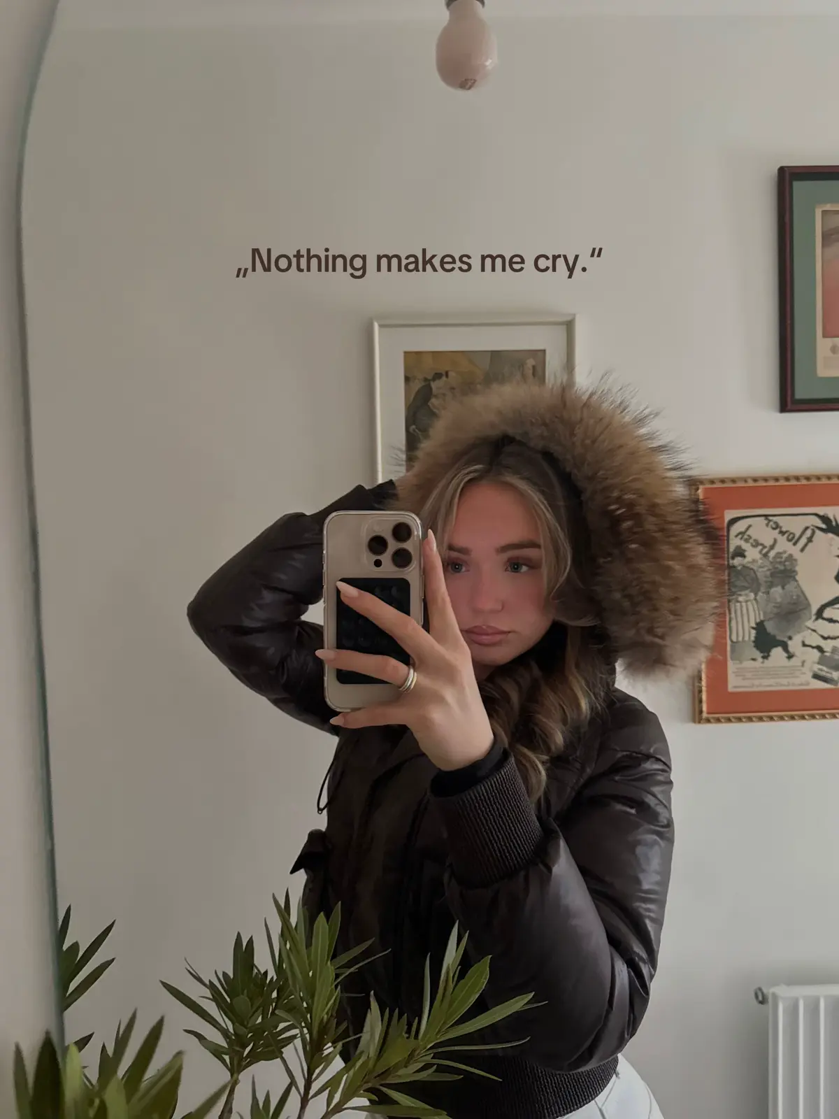 A well-lit mirror selfie featuring a woman holding up a phone, partially obscuring her face. She's wearing a dark hooded jacket with fur trim. The background includes framed artwork and a light fixture, all in neutral tones.