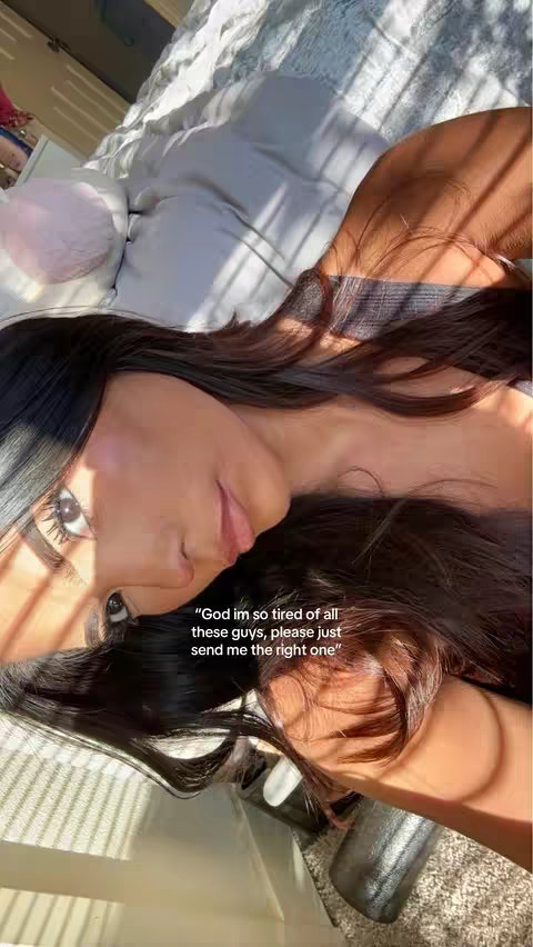 The cover image features a young woman with long dark hair, lying in bed. She's holding the camera and looking at it with a slight smile, the top of her hair casting a shadow over her face. Text overlayed on the bottom of the image reads, "God im so tired of all these guys, please just send me the right one."