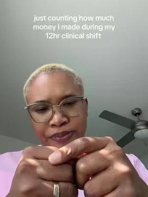A woman with short blonde hair, wearing glasses and a pink shirt. The woman is holding her hands in front of her, fists close together. The background is a bit out of focus, with a ceiling fan visible.