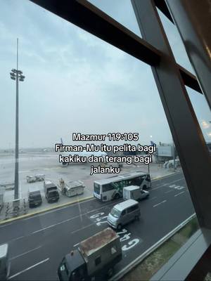 An overcast day view from inside a building, likely an airport. The scene includes a runway with parked vehicles and buses and a text overlay containing a Bible verse.