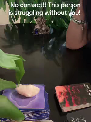 The image shows a dark, reflective surface with various objects arranged on it. In the center, there is a stack of tarot cards with a blurred image on top. To the left, there's a pile of purple cards topped with a white stone. A green leafy plant is placed near the background. The upper-right corner captures a leg and part of an arm, along with a wrist adorned with a bracelet. Text in a white font is overlaid on the top-left, saying 'No contact!! This person is struggling without you!'