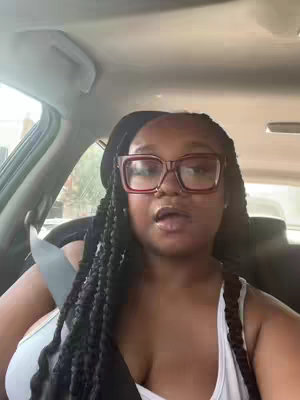 The image displays a woman in a car, likely filming herself, with a focus on her face. She is wearing large-framed glasses and has her hair styled in braids. The car's interior is visible, providing a background.