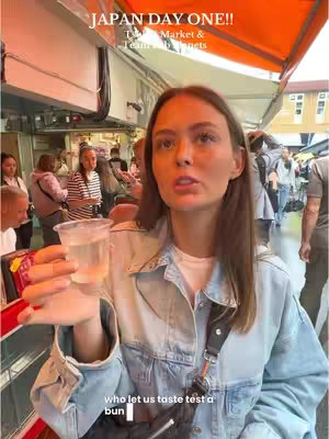 A woman in a denim jacket is holding a clear cup with a liquid inside. Background includes other people, market stalls, and a sign that says 'Japan Day One.'