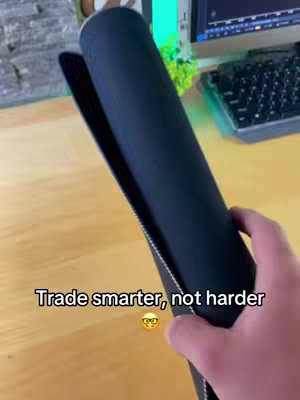 A hand holds a rolled-up, black mousepad, revealing a glimpse of a desk with a computer and keyboard in the background. The lighting appears neutral, and the composition focuses on showcasing the product.