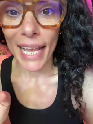 A close-up shot of a woman's face, with a focus on her eyes and mouth. She wears large, square-framed glasses. Her dark curly hair frames her face, and she appears to be speaking. The background is blurred, with a hint of pink.