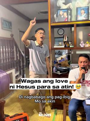 The photo shows two men in a living room setting. One man is standing with his arm raised, singing or speaking energetically. The other man is seated, holding a microphone and also appears to be singing. Text overlays are present, suggesting this is a cover song or worship video.