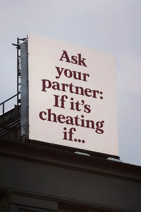 A photograph shows a large billboard mounted atop a building, set against an overcast sky. The billboard has a white background with text in a deep red or burgundy color. It reads, 'Ask your partner: If it's cheating if...' The text is arranged in three lines to create a visual emphasis on the question.