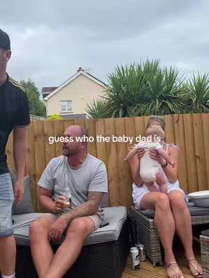 An outdoor scene featuring three adults and a baby. The focus is on a man seated, a woman holding a baby, and a third person. Text overlaying the image asks 'guess who the baby dad is'. The scene is in a patio setting with a wooden fence background.