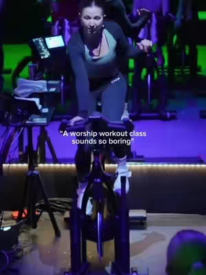 The cover image shows a woman riding a stationary bike in a dimly lit cycling studio. She is wearing athletic attire and a headset microphone. The text overlay reads "A worship workout class sounds so boring". The background has other cyclists and green and purple lighting.