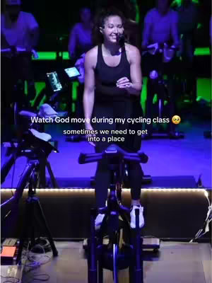 A woman, probably in her 20s or 30s, is standing on a stationary bike in a cycling studio. She is smiling and looking towards the camera. Text overlays the image, conveying a message. The background is blurred, showing other people on stationary bikes in a studio lit with blue and green lights.