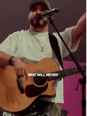 A man wearing a baseball cap, a light green t-shirt, and a cross necklace is playing a wooden acoustic guitar and singing into a microphone. Text on image: WHO WILL NEVER.