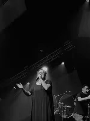 Monochrome image of a woman singing on stage with a microphone. She is wearing a dark dress, and other band members are in the background. Stage lighting is focused on the singer. The image is mostly black and white.