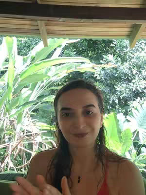 A woman is seen in front of a lush green background, likely outdoors. She is looking directly at the camera with a slight smile, wearing a red top and a small necklace. Her long hair is down. Her hands are slightly raised in front of her. The background is blurred, but leafy plants and glimpses of a structure are visible. The overall composition suggests a casual and natural setting.