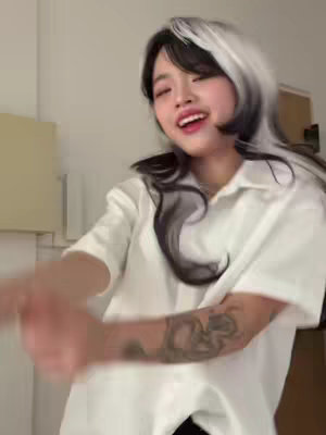 A person with black and white hair smiles at the camera while dancing in front of a white wall. They are wearing a white button-down shirt and have tattoos on their arm.