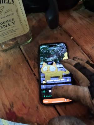 A person's hand holding a phone displaying a cartoon cat illustration. The phone is on top of a wooden surface, with a bottle of Jack Daniels visible in the background. The hand appears to be tattooed.