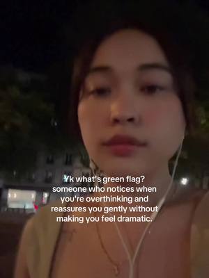 A selfie-style shot of a woman, framed from the chest up, taken at night. She's looking towards the camera with a subtle expression. The background is blurred, featuring indistinct shapes and dark foliage, with hints of streetlights. She is wearing earbuds, suggesting she's listening to something, and a simple necklace. Overlayed text in a light sans-serif font poses a question in the upper-left area about "green flags" and responds with a description about someone who notices overthinking and reassures gently.