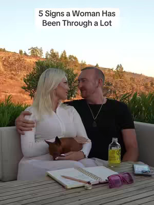 The cover image features a blonde woman and a man with reddish-brown hair sitting together at a table outdoors. The woman is holding a small dog. A notebook, pencil, sunglasses, and a bottle are on the table. The background shows a hillside with trees under a bright sky.