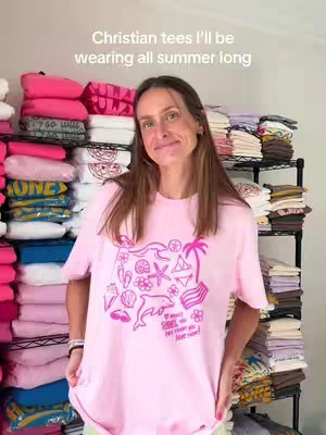 A woman stands in a room filled with stacks of folded clothes. She's wearing a pink t-shirt with various beach-themed graphics. The text overlay reads, 'Christian tees I'll be wearing all summer long'.
