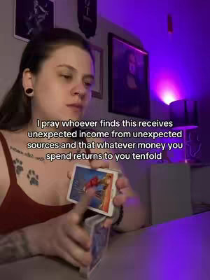 A medium shot captures a woman holding up a deck of tarot cards. She is facing toward the camera with a neutral expression. The background shows wall art and a lamp to the right. The lighting is dim and colored, appearing purple.