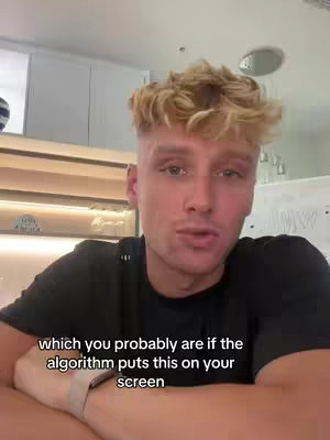 A young man with styled blonde hair is centered in the frame, looking directly at the viewer. He's wearing a black t-shirt and has a light-colored watch. The background is a blurred interior with neutral colors, partially visible cabinets and a light fixture.