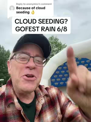 A man wearing a black cap, glasses, and a plaid shirt is pointing upwards with his index finger. The background includes a cloudy sky and a white and blue structure, along with some greenery.
