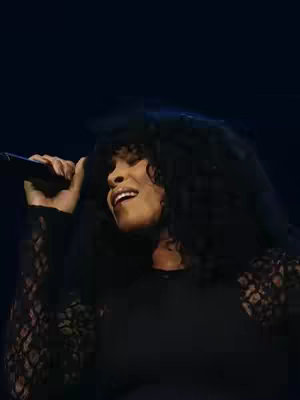 A woman with dark curly hair is singing into a microphone on a dark stage. She wears a black long-sleeved top with lace detailing on the sleeves. The background is a solid dark blue. The image is lit from the front, illuminating the woman's face and upper body.
