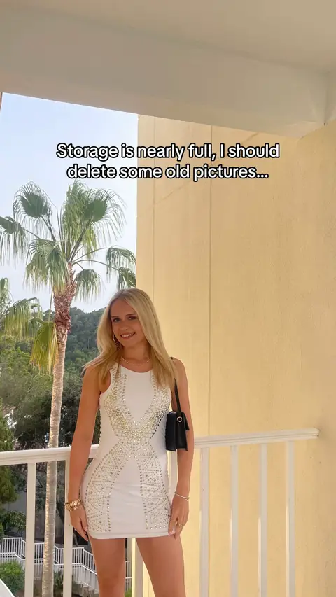 A woman with long blonde hair stands on a balcony. She wears a white dress with a decorative pattern and a black purse. The background includes a building and palm trees. Text overlay says "Storage is nearly full, I should delete some old pictures...".