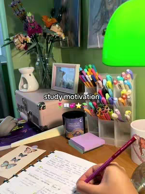 A top-down shot of a desk setup with a notebook open. There's a pen in the hand of the person and the hand is in frame with the pen on the notebook. The desk is full of study supplies, including colourful pens, a candle, post-it notes and decorations like flowers and a framed picture. The color pallette is diverse.