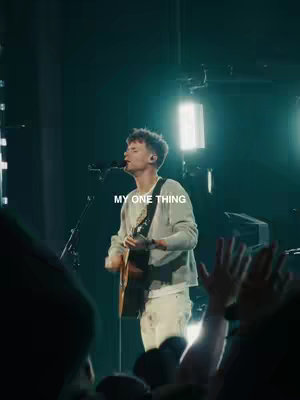 A live music performance captured in a vertical frame. A man stands center frame, playing an acoustic guitar and singing into a microphone. He's wearing a light-colored sweater and pants, with his hair styled up. Soft stage lighting bathes the scene, with some brighter lights in the background. The foreground includes silhouettes of audience members and raised hands. Text overlayed says, "MY ONE THING."