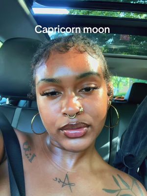 A selfie taken inside a car. The main subject is a person with dark skin, wearing gold hoop earrings and a septum ring. They have long eyelashes, dark eyebrows, and full lips. A tattoo of a star is visible on their chest. The background shows the interior of a car and a view of the outside through the sunroof.