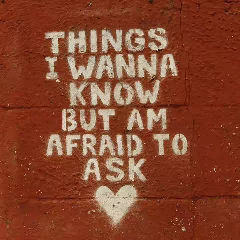 The image is a close-up of a weathered, reddish-brown wall. Stenciled onto the wall is white text that reads: "THINGS I WANNA KNOW BUT AM AFRAID TO ASK," followed by a simple white heart shape at the bottom center. The text is arranged in three main lines, with the heart slightly below. The paint appears slightly faded and textured, giving it a rough, handmade feel. The overall composition is simple, with the text and heart being the central focus against the textured backdrop.