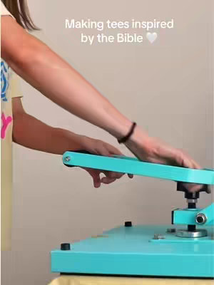 A person is shown from the elbows down, pressing a large, teal heat press. The person's arms and hands are visible, with a black bracelet on the wrist. The person is pressing a teal machine; a light cream-colored background is visible. Text overlays at the top: 'Making tees inspired by the Bible' and a white heart emoji.