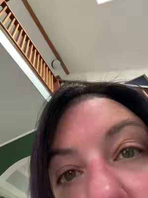 A close-up shot of a woman's face, seen from the forehead down, with her eyes as the main focus. The background is the corner of a room with a staircase. The lighting is natural, and the overall feel is casual.