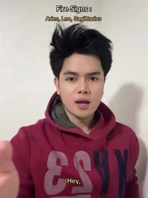 A young man with dark hair, wearing a red hoodie, appears in a casual selfie video. The overlaid text specifies "Fire Signs: Aries, Leo, Sagittarius" at the top, and a smaller text bubble contains the word "Hey," near the bottom. The composition is front-facing, capturing the man's upper body against a neutral, cream-colored background