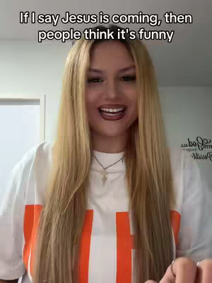 A young woman with long blonde hair, wearing a white shirt with an orange stripe, smiles widely at the camera. The image is a selfie taken in a well-lit room, with text overlaid at the top. In the background, a door and some writing on the wall are visible.