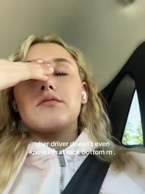 A young woman is seated inside a car, the interior is visible. She has blonde hair, and is wearing a light-colored hoodie. The woman is covering her eyes with her left hand. The background is the car interior. Below the image, the text overlay reads: "uber driver doesn't even know i'm at rock bottom rn."